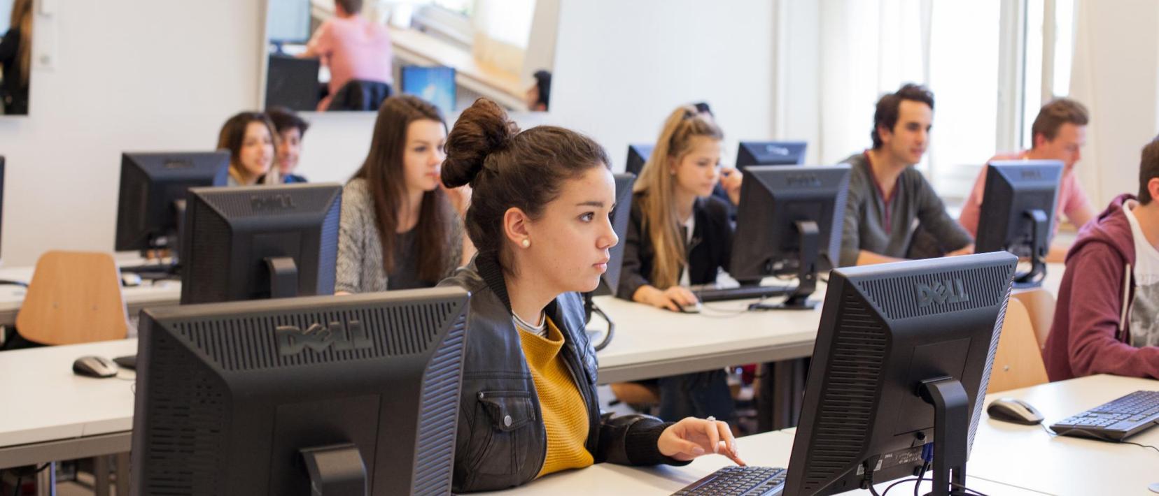 Blick in ein Klassenzimmer, in dem Schülerinnen und Schüler an Computerbildschirmen arbeiten.