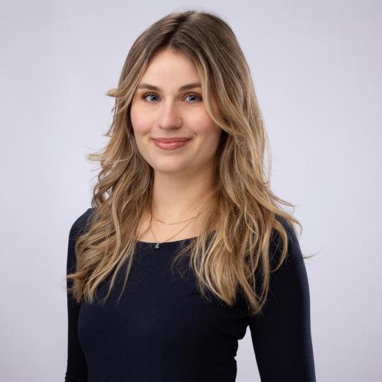 Sarah trägt ein schwarzes Oberteil. Sie hat lange blonde Haare und lächelt freundlich. 