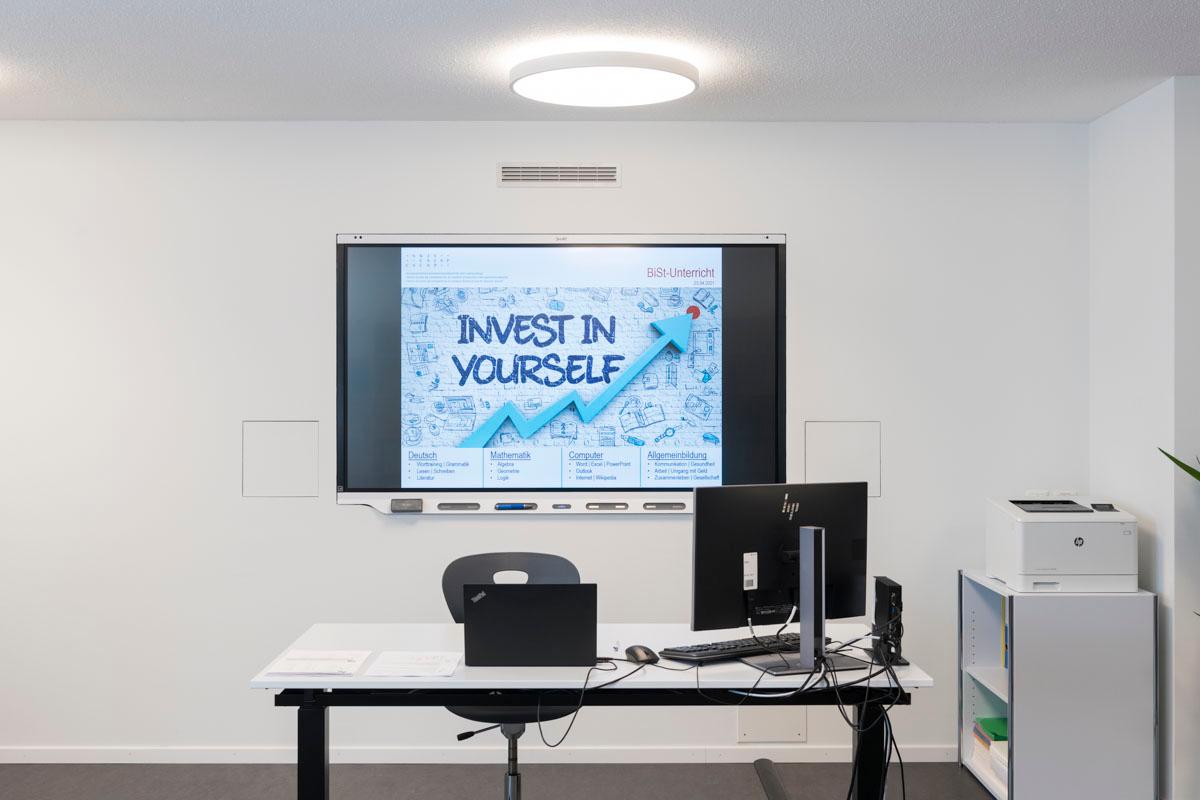 Weisser Büroschreibtisch mit Stuhl und Bildschirm. An der Wand hängt ein grosser Bildschirm, auf dem ein Bild sagt «Invest in yourself».