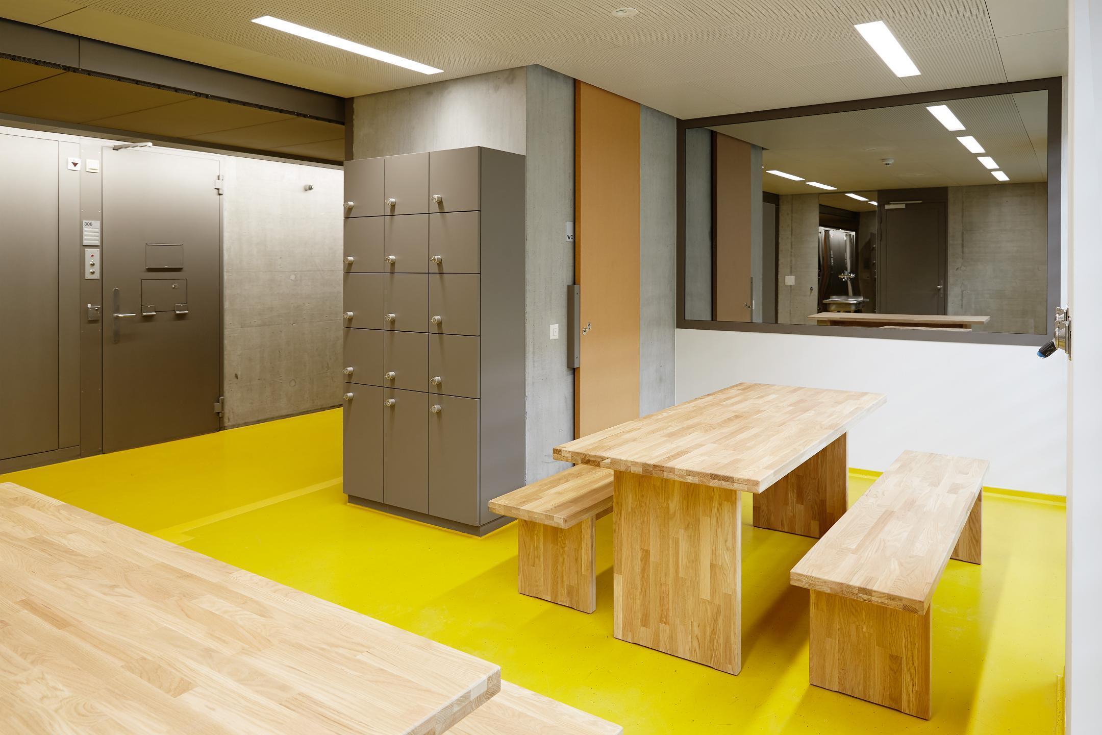 Hell beleuchteter kahler Raum mit Holztisch, Bänken, Spiegelscheibe und Einwegspiegel