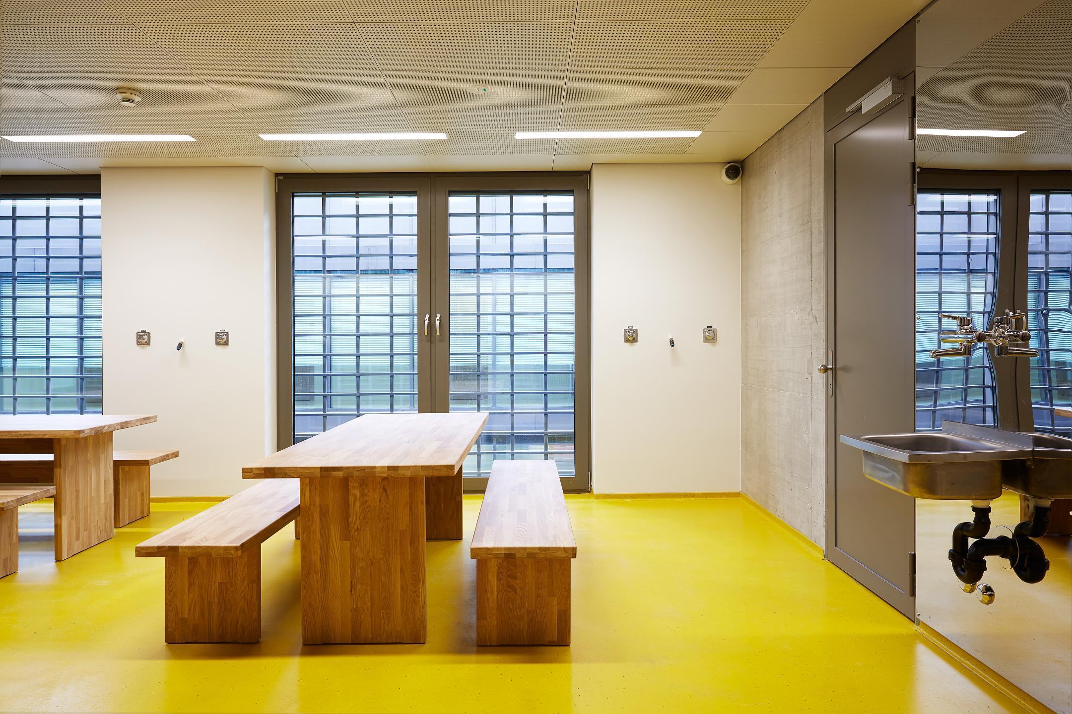 Hell beleuchteter Raum mit Tischen und Bänken vor vergitterten Fenstern