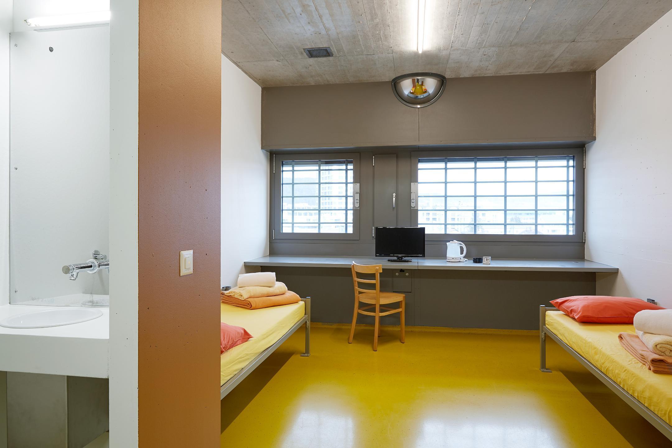 Raum mit zwei Betten, Fernseher, Lavabo und vergitterten Fenstern