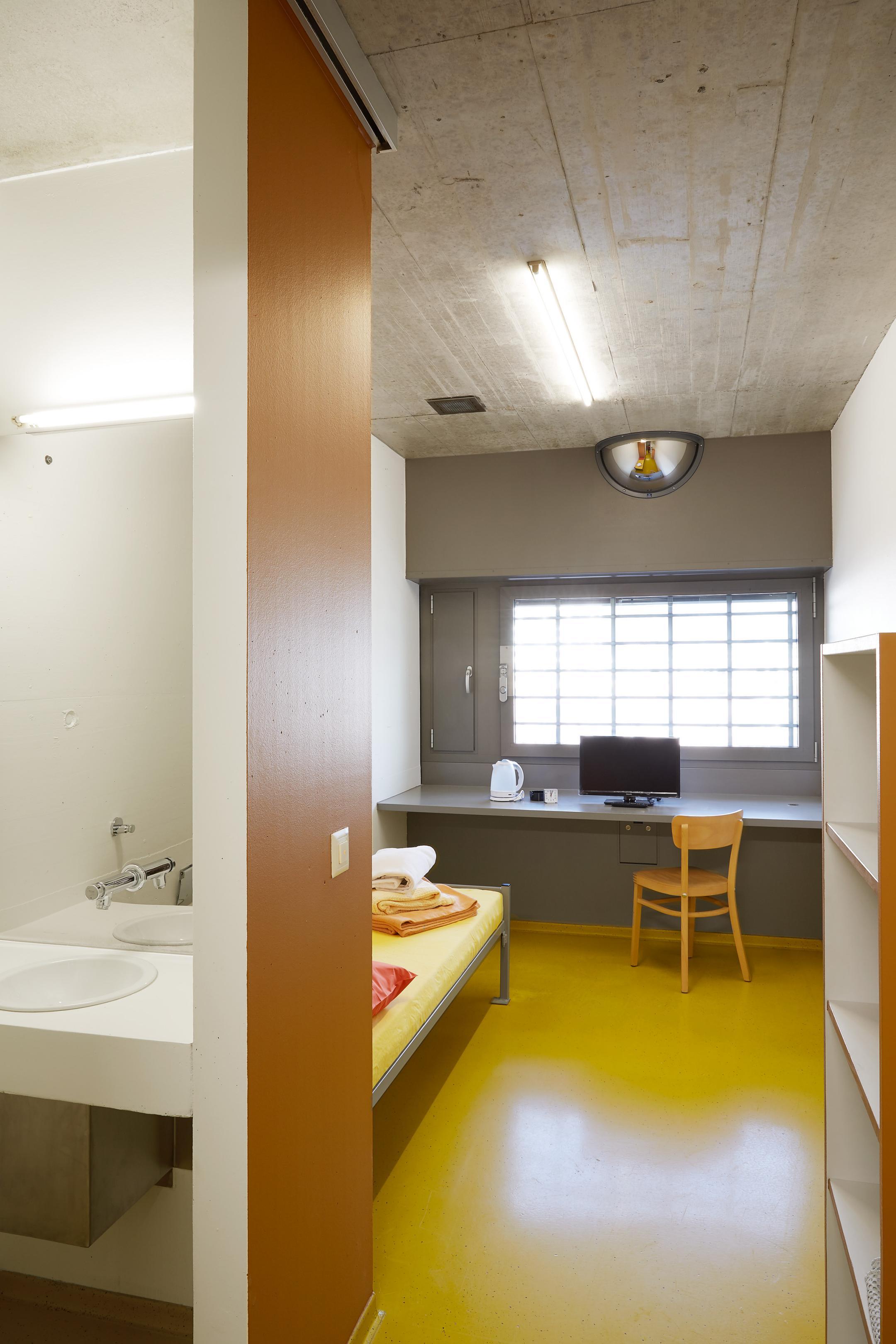 Raum mit Bett, Fernseher, Lavabo und vergitterten Fenstern