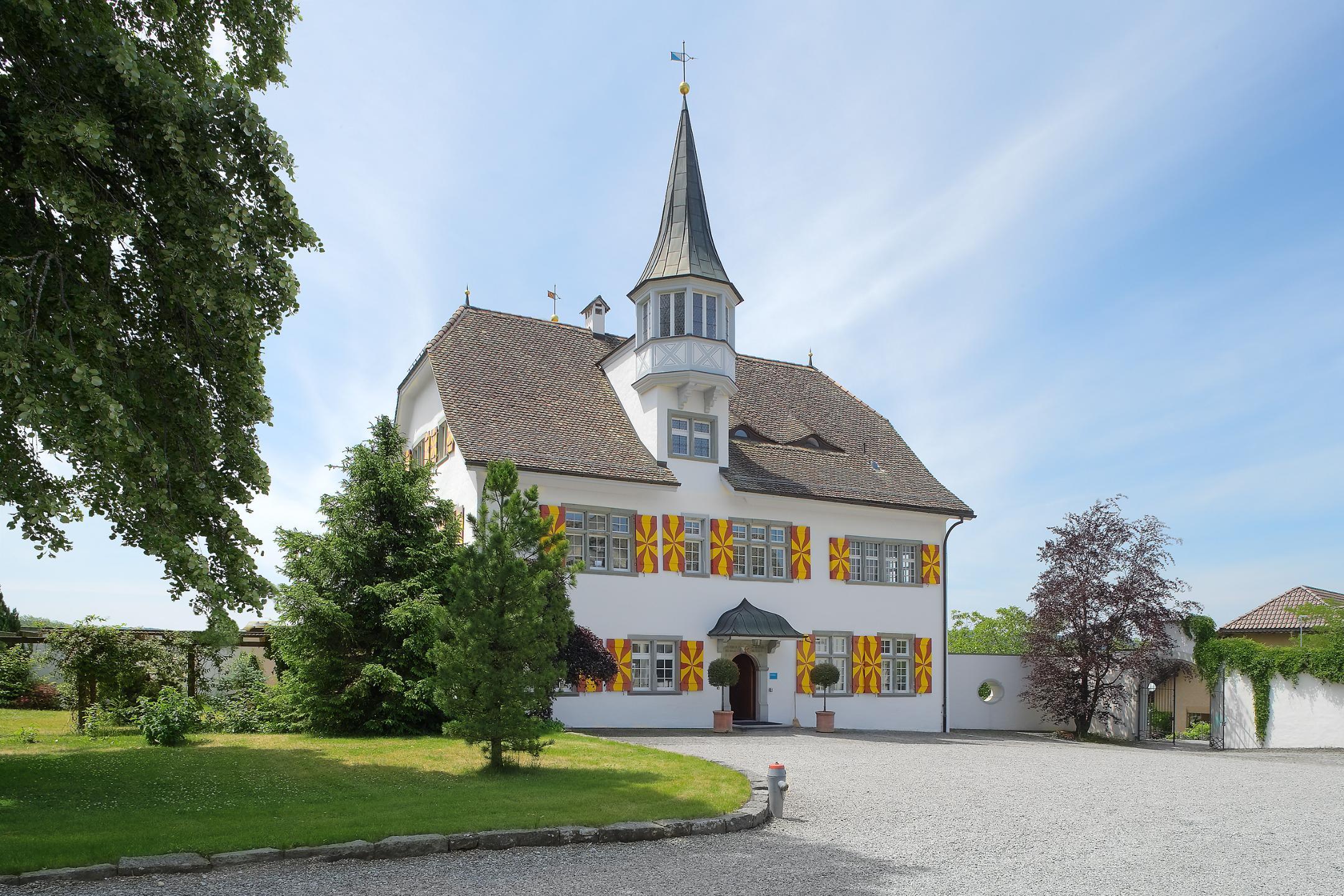 Gebäude mit gelben Fensterläden vor Kiesplatz und angrenzender Wiese.