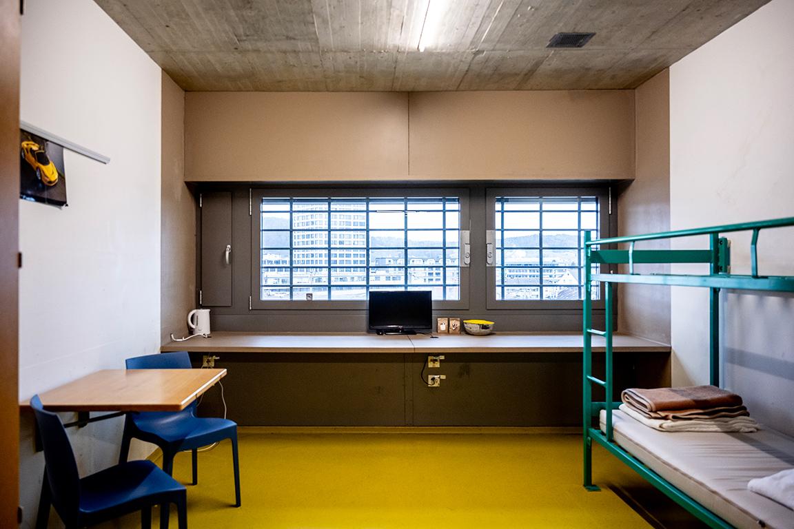 Eine Zelle mit gelbem Boden, einem Tisch mit zwei Stühlen und einem Etagenbett.