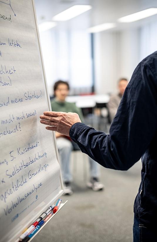 Der Kursleiter zeigt mit seiner Hand aus etwas auf dem Flipchart.