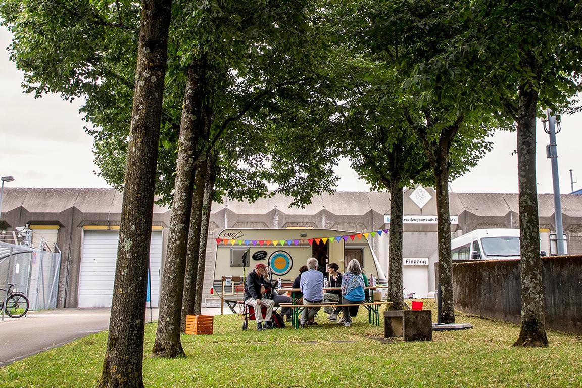 Personen sitzen an einem Tisch vor einem Wohnwagen. Der Wohnwagen steht vor dem Gefängniseingang.