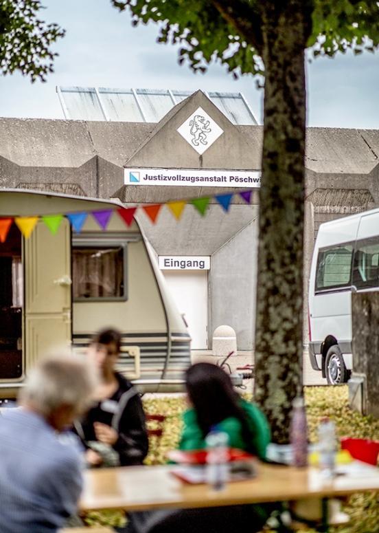 Im Fokus ist der Eingang der JVA Pöschwies. Im verschwommenen Vordergrund des Fotos sieht man den Wohnwagen und 3 Personen, die am Tisch davor sitzen.
