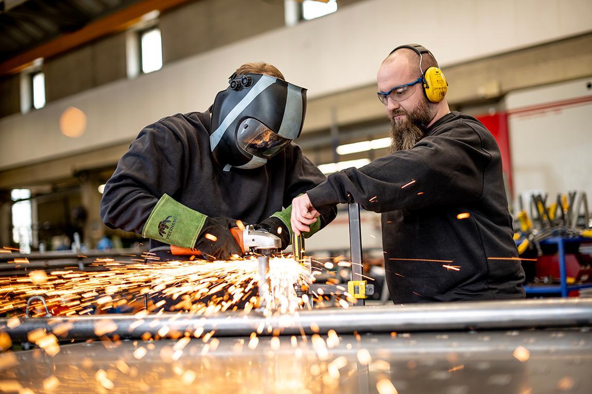 Ein Mitarbeiter und ein straffälliger Jugendlicher arbeiten zusammen an Metall, es fliegen Funken.