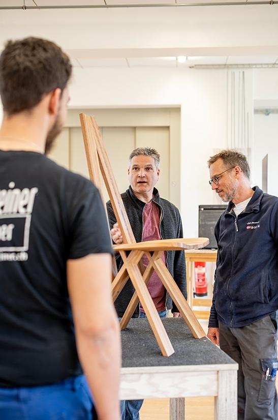 Zwei Männer stehen in der Schreinerei und besprechen mit einem jungen straffälligen Mann einen Stuhl aus Holz.