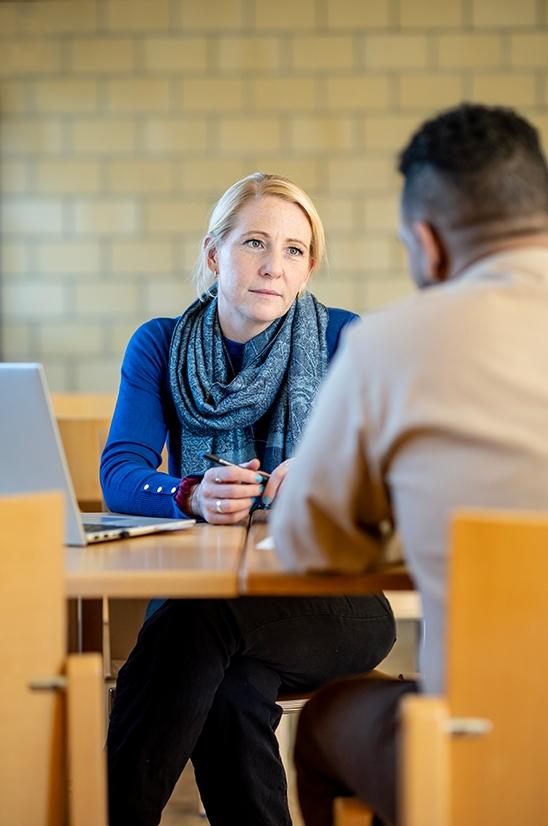 Sozialdienstmitarbeiterin beratet einen Inhaftierten im Besuchsraum. Sie sitzen am Tisch.