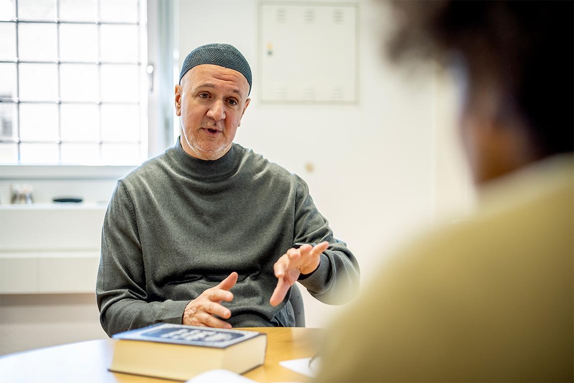 Der muslimische Seelsorger sitzt am Tisch. Auf dem Tisch liegt der Koran und er spricht mit einem Inhaftierten.
