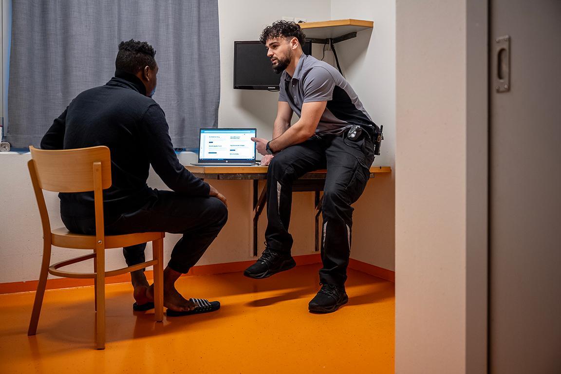 Ein inhaftierter Mann sitzt am Tisch im Haftraum vor einem Laptop. Ein Mitarbeiter spricht mit ihm und erklärt ihm das Programm auf dem Laptop.