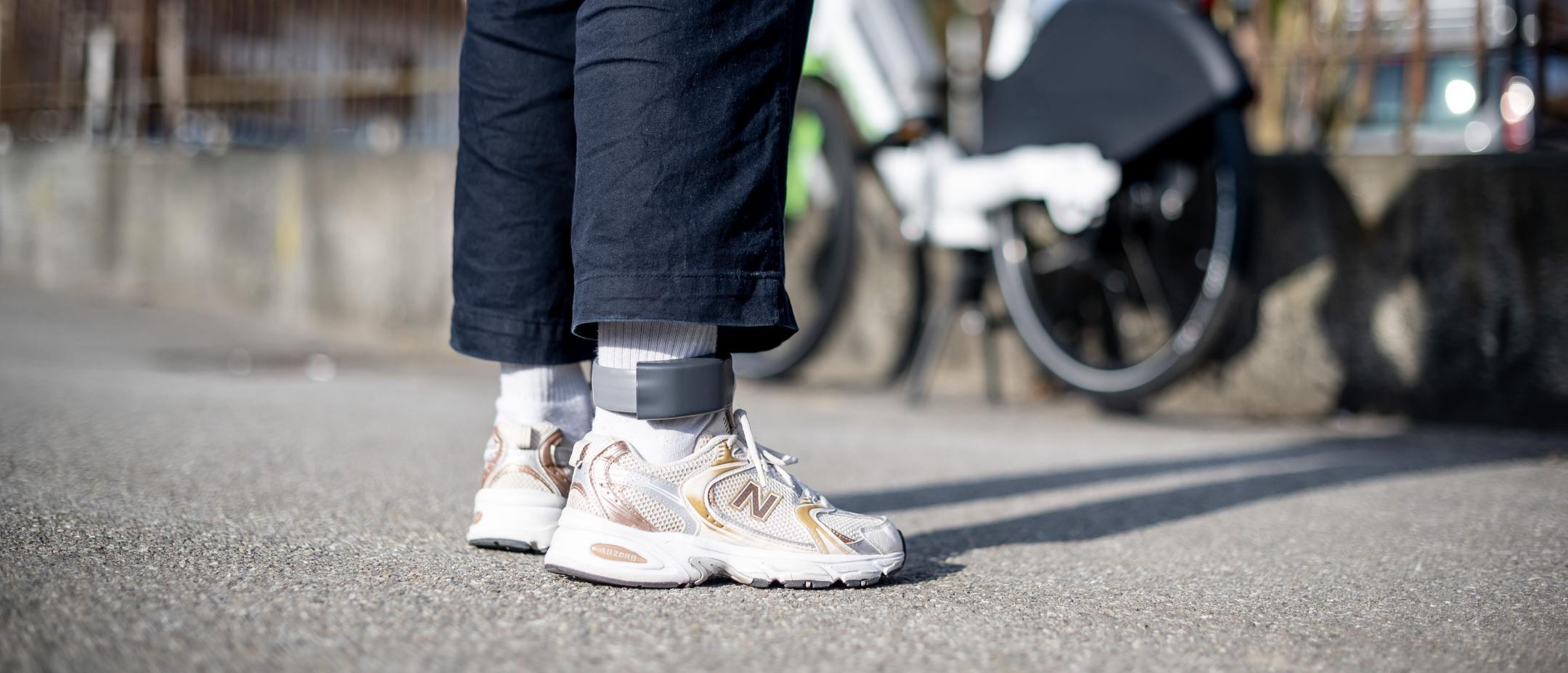 Eine Person steht vor einem Fahrrad und trägt eine elektronische Fussfessel um den Knöchel.