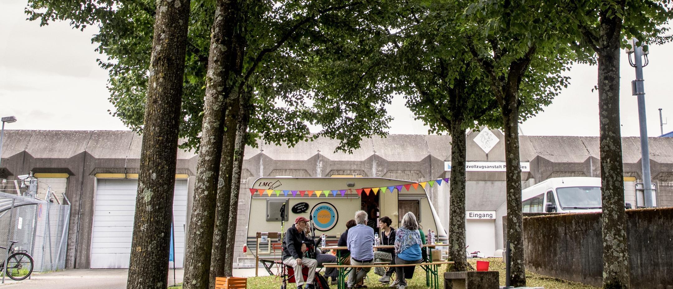 Menschen sitzen an einem Tisch vor einem Wohnwagen, dieser steht wiederum vor dem Eingang der JVA Pöschwies.