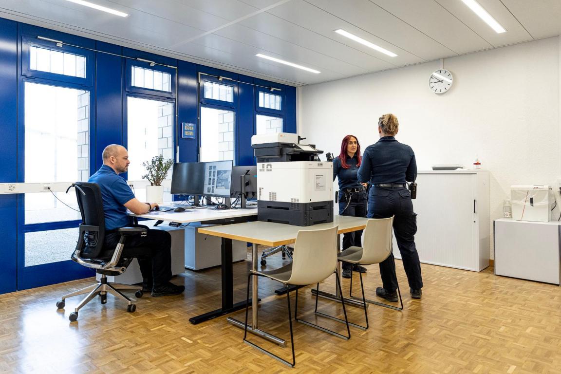 In einem Büro sitzt ein Mann am Tisch und arbeitet. Ihm gegenüber stehen zwei Frauen und unterhalten sich.