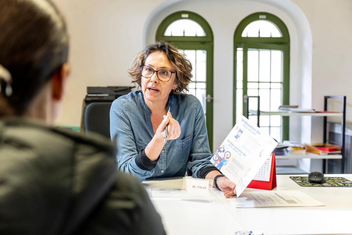 Eine Frau sitzt im Büro. Ihr gegenüber sitzt eine andere Person, deren Gesicht man nicht sieht. Die Frau hält eine Broschüre und einen Stift in der Hand und spricht erklärend mit der anderen Person.