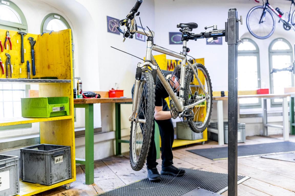 Ein Velo hängt an einer Vorrichtung in der Luft, während ein Mann es mit Werkzeug in der Hand repariert.