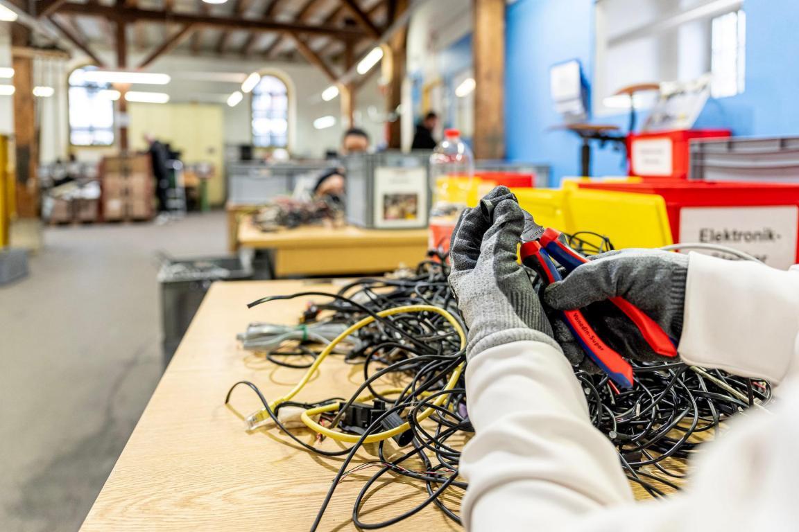 Eine Person sitzt am Tisch und hält eine Zange und Kabel in der Hand, im Hintergrund ist eine Werkstatt.