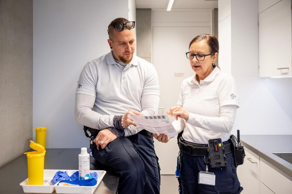 Ein Mann und eine Frau stehen in einer Küche. Beide tragen Arbeitsuniform. Sie unterhalten sich und schauen gemeinsam auf einen Zettel.