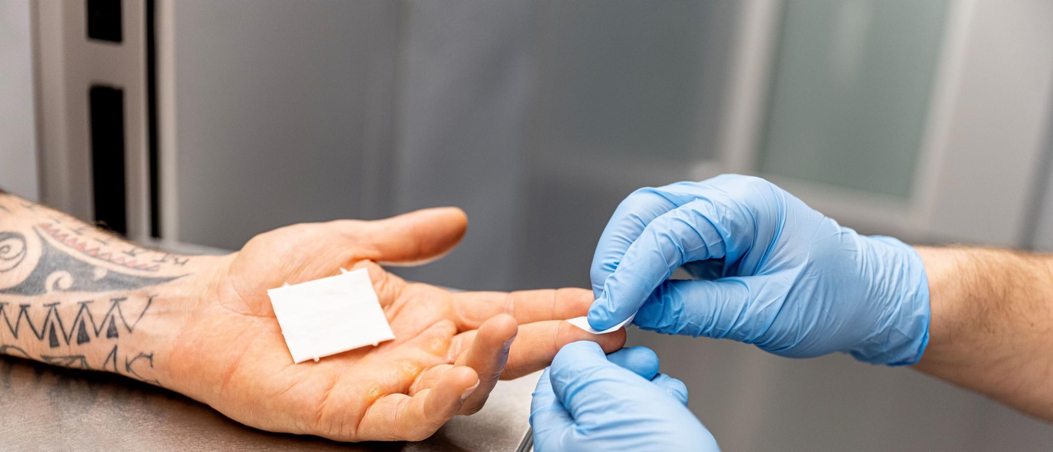 Einer inhaftierten Person wird durch die Zellenklappe am Finger Blut abgenommen. Die Person, die Blut abnimmt, trägt blaue Gummihandschuhe.