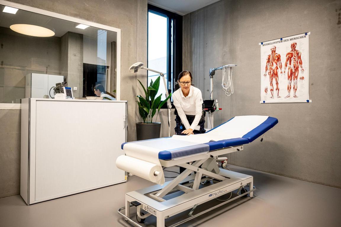 In einem Behandlungszimmer steht eine blaue Liege mit Hygieneschutz. Eine Mitarbeiterin streicht diesen Schutz gerade glatt.