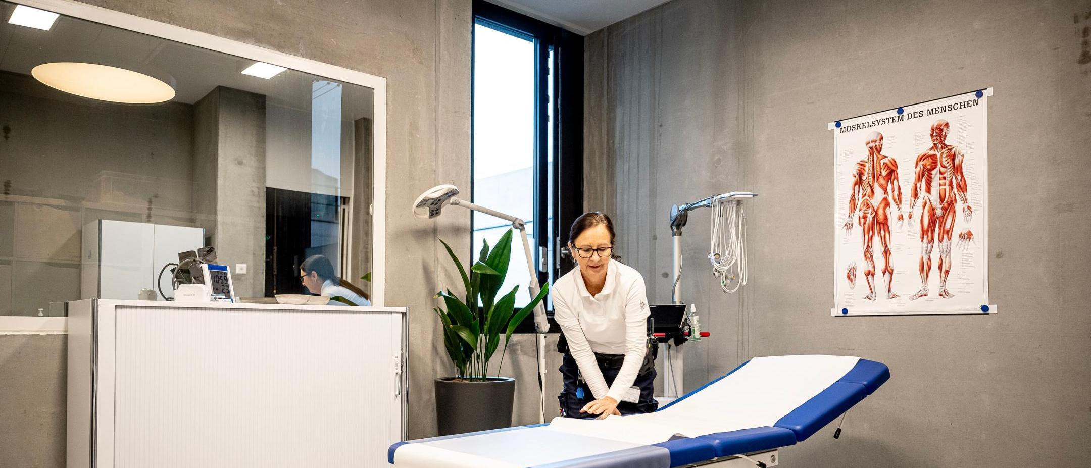 In einem Behandlungszimmer steht eine blaue Liege mit Hygieneschutz. Eine Mitarbeiterin streicht diesen Schutz gerade glatt.