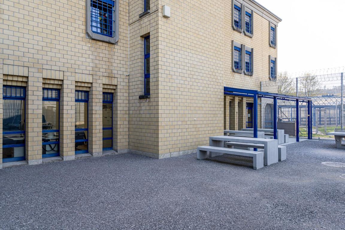 Backsteingebäude mit Kiesplatz davor. Auf dem Kiesplatz stehen Tische aus Beton, die von einer blauen Pergola überdacht sind.