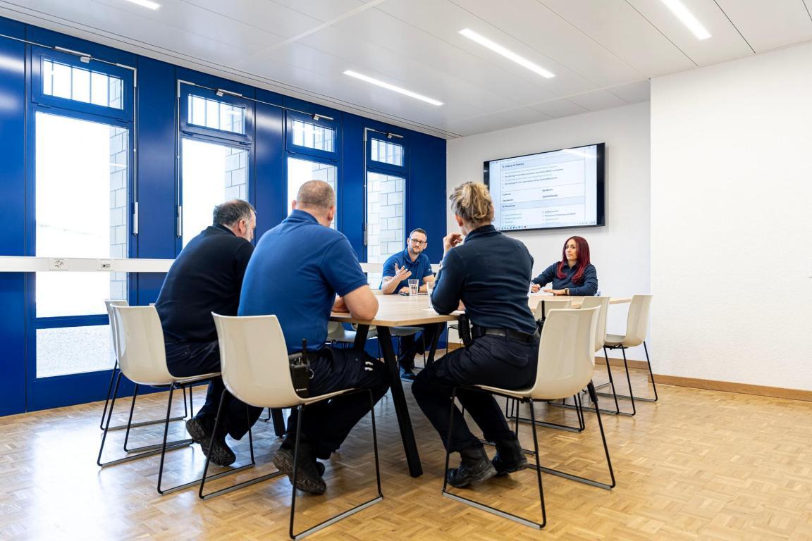 Fünf Personen sitzen in einem Sitzungszimmer an einem Tisch und schauen auf einen Bildschirm an der Wand. Sie diskutieren..