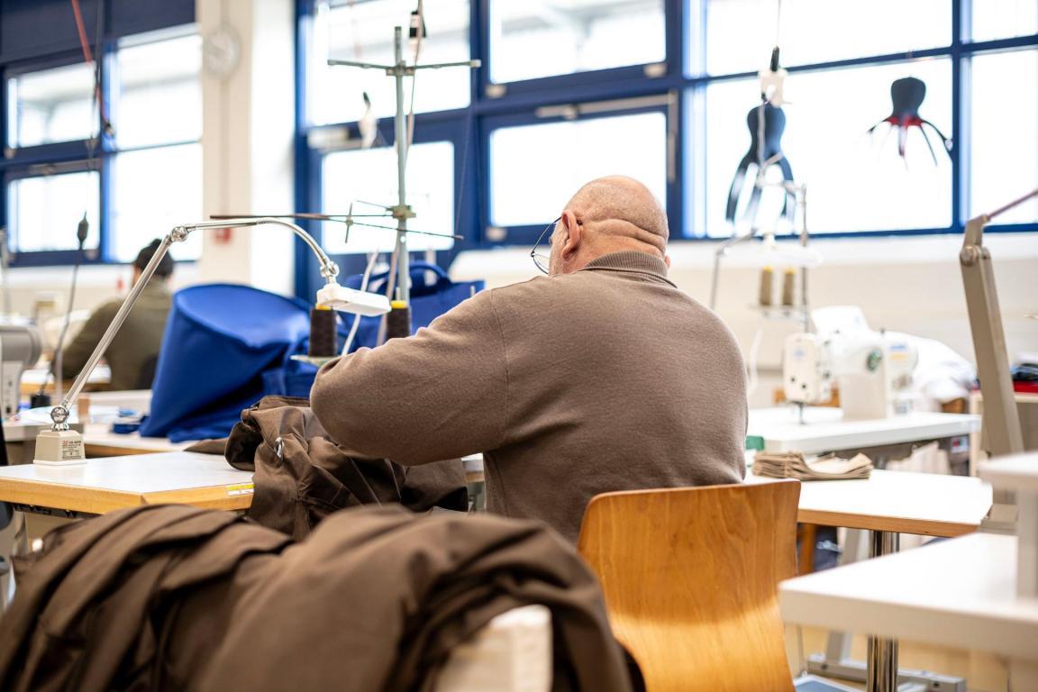 In einer Näherei sitzt ein Mann mit Glatze an einem Tisch und näht. Er trägt einen braunen Pullover.