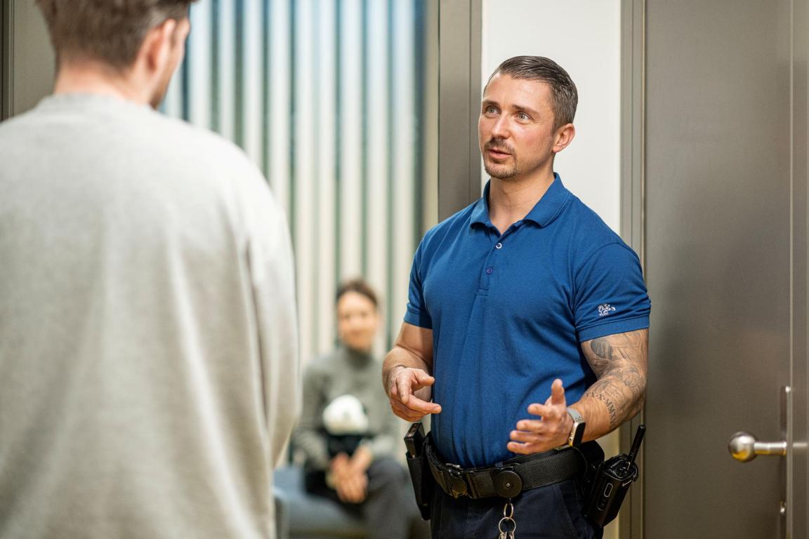 Mitarbeiter des Gefängnis weist einen inhaftierten Mann seinem Besuch zu. Der Besuch wartet im Zimmer im Hintergrund.