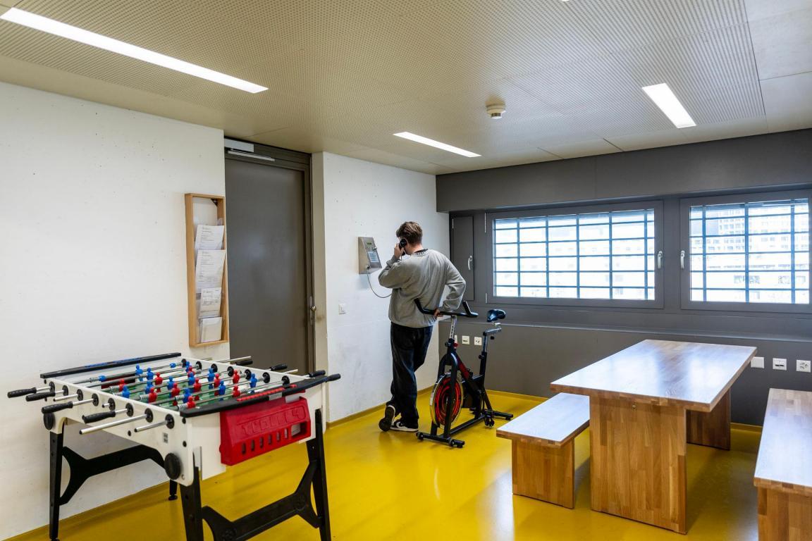 Ein Mann telefoniert an einem Wandtelefon, während er sich an ein Indoorfahrrad lehnt. Neben ihm stehen ein Tisch und ein Tischfussball.