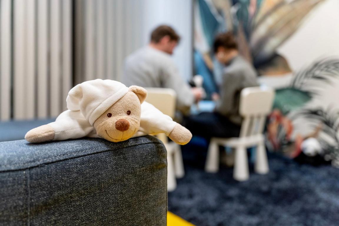 Ein Plüschtier liegt auf einem Sofa. Im Hintergrund sitzen ein Mann und eine Frau an einem Tisch für Kinder und unterhalten sich.