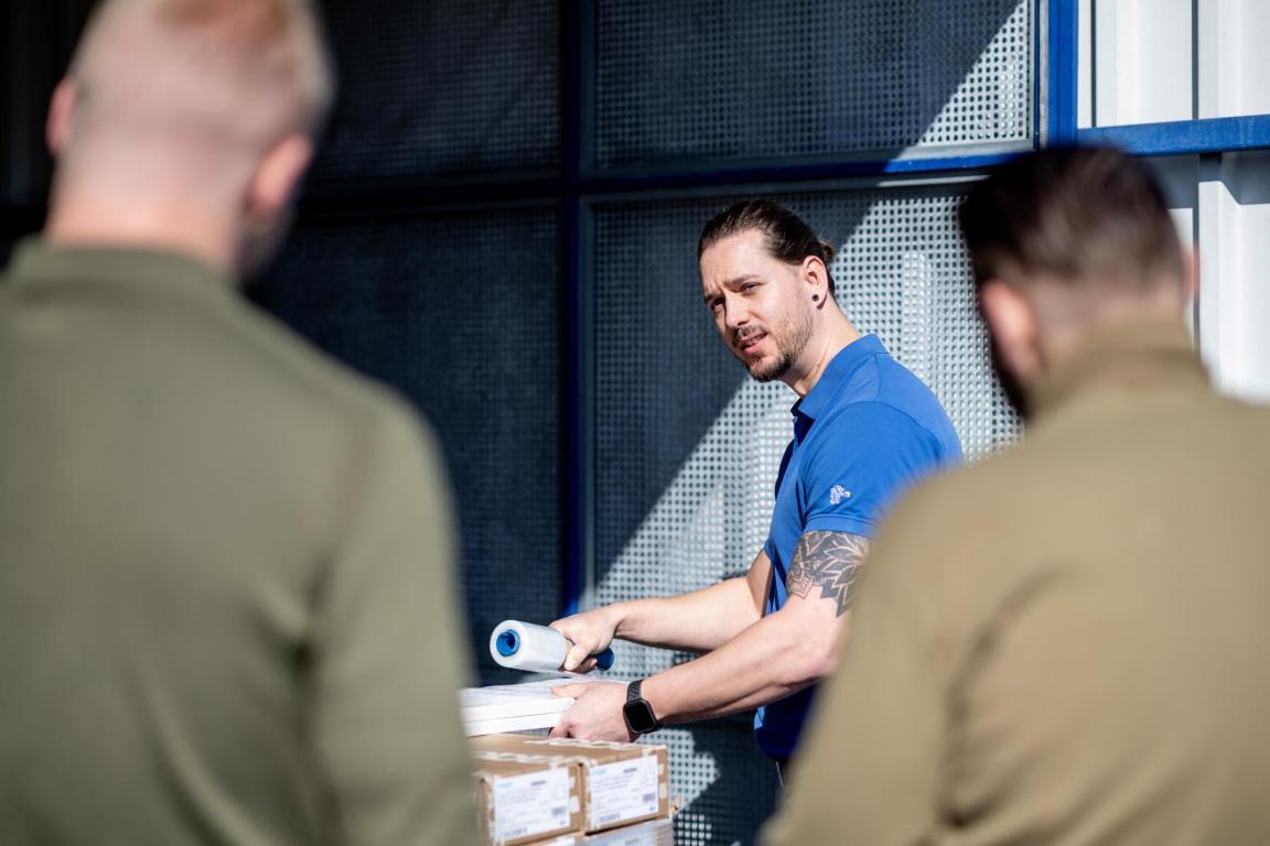 Mitarbeiter zeigt zwei inhaftierten Männern, die mit dem Rücken zur Kamera stehen, wie man etwas in Folie einwickelt.