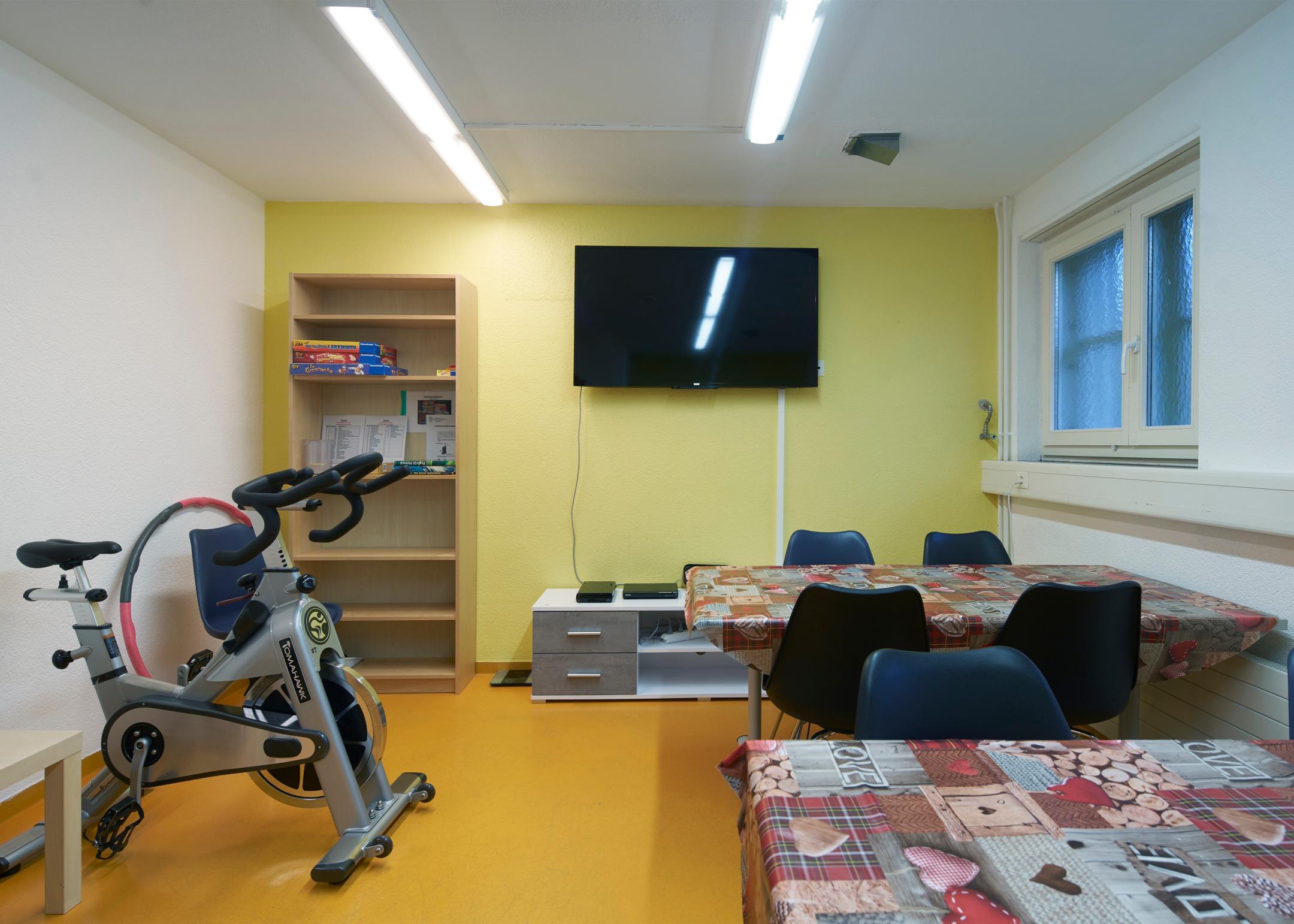 Auf dem Bild zu sehen sind ein Bücherregal mit Brettspielen, diverse Sportgeräte, Tische, Bestuhlung und ein Fernseher.
