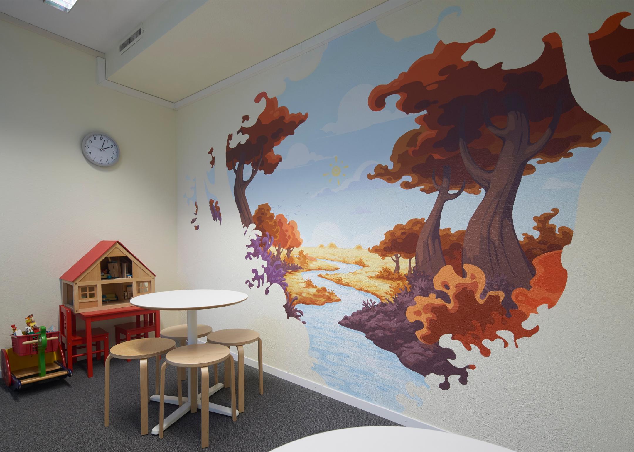 Tisch und Bestuhlung und diverse Kinderspielzeuge, unter anderem ein Puppenhaus. Diese stehen vor einer farbigen, mit der Illustration einer Naturlandschaft verzierten Wand.