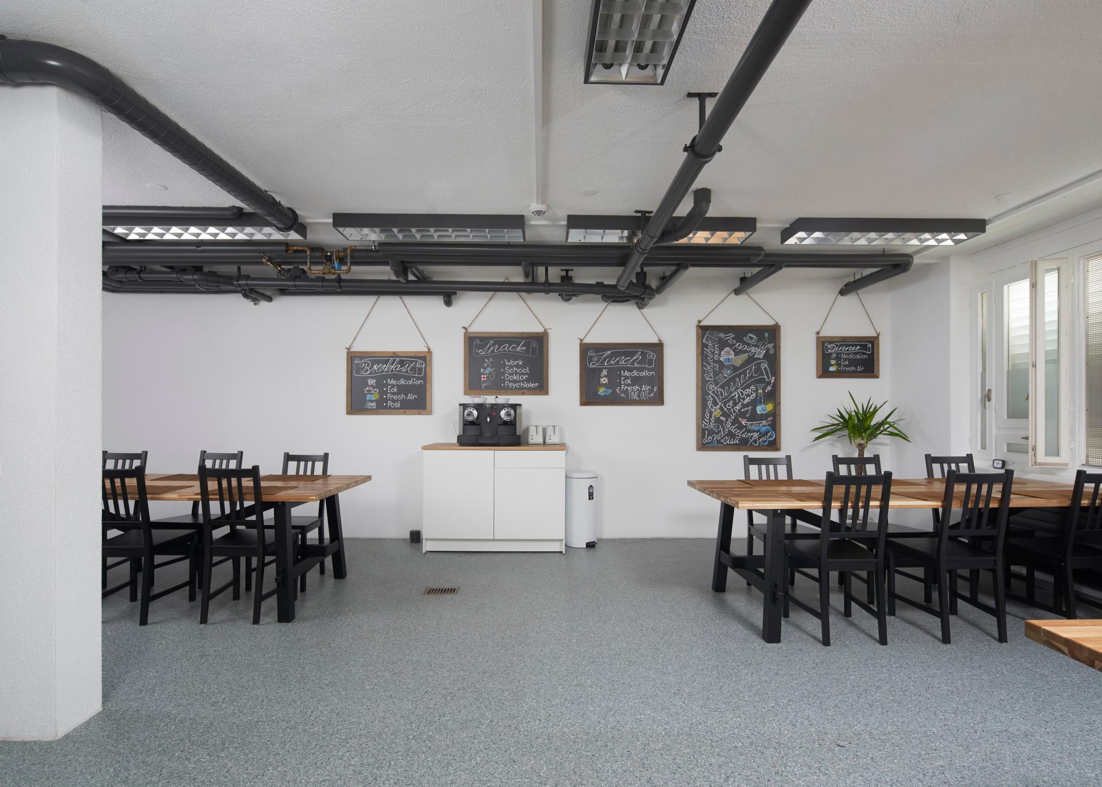 Auf dem Bild zu sehen sind Tische, Stühle, eine Kaffeemaschine und verschiedene Wandtafeln mit farbigen Illustrationen.