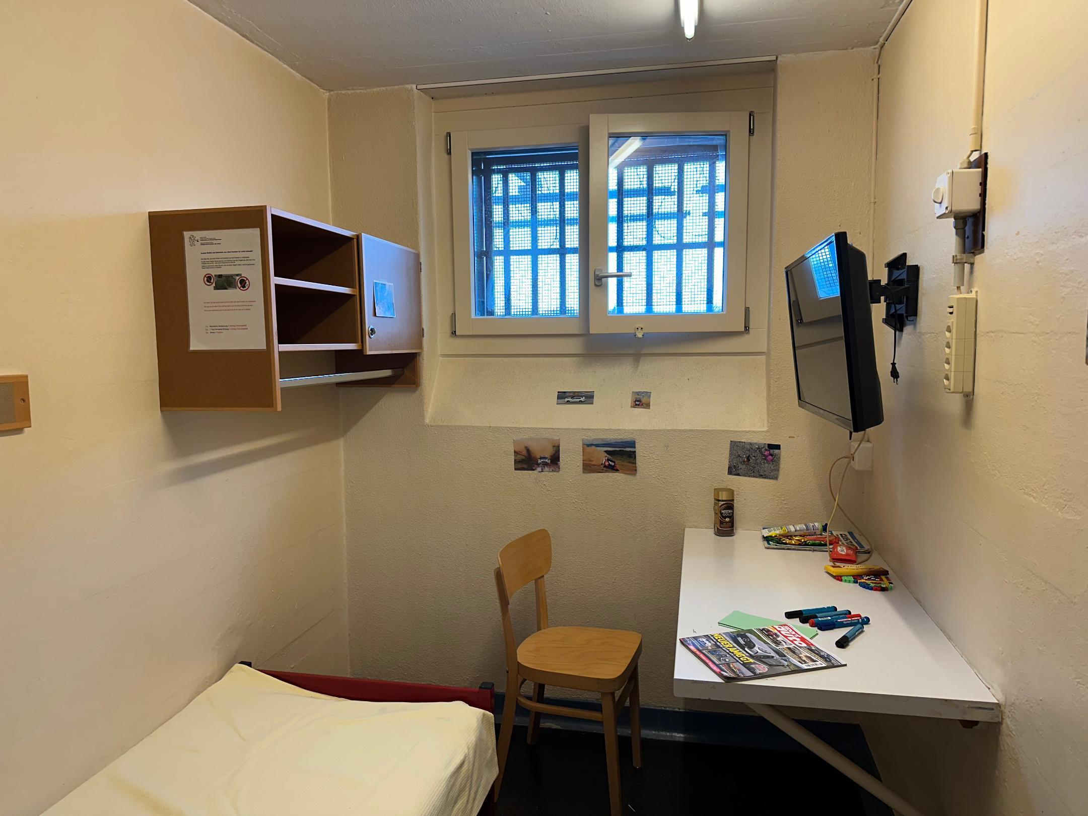 In der Zelle ist ein Bett, ein Tisch mit einem Stuhl, ein Fenster und ein Fernseher.