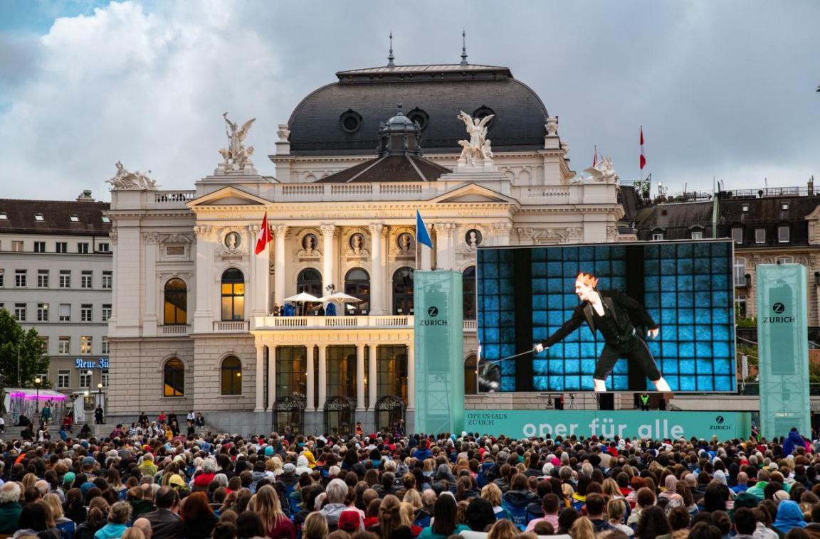 Vor dem Opernhaus Zürich verfolgt eine grosse Menschenmenge eine Aufführung auf einer Grossbildleinwand. Die Veranstaltung «Ballett für Alle 2019» findet unter freiem Himmel statt. Das Opernhaus steht im Hintergrund und bildet eine eindrucksvolle Kulisse für den Anlass.