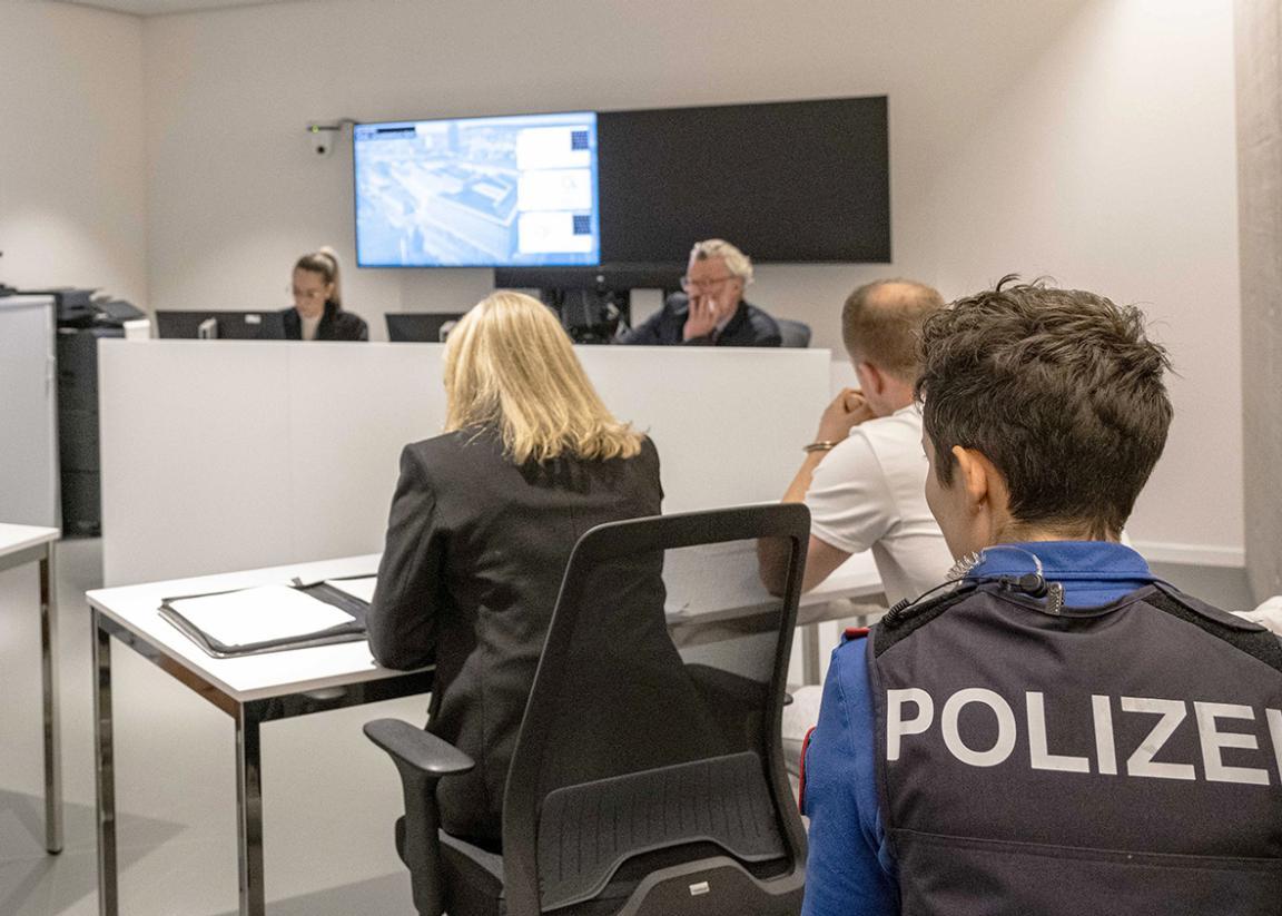Mehrere Personen sitzen in einem Einvernahmeraum. Im Vordergrund sitzt eine Polizistin mit dem Rücken zur Kamera, während vorne eine Einvernahme stattfindet.