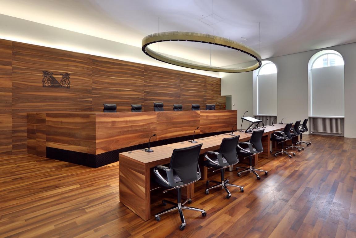 Ein leerer Gerichtssaal mit einer erhöhten Richterbank aus Holz, mehreren Mikrofonen und Sitzplätzen davor.