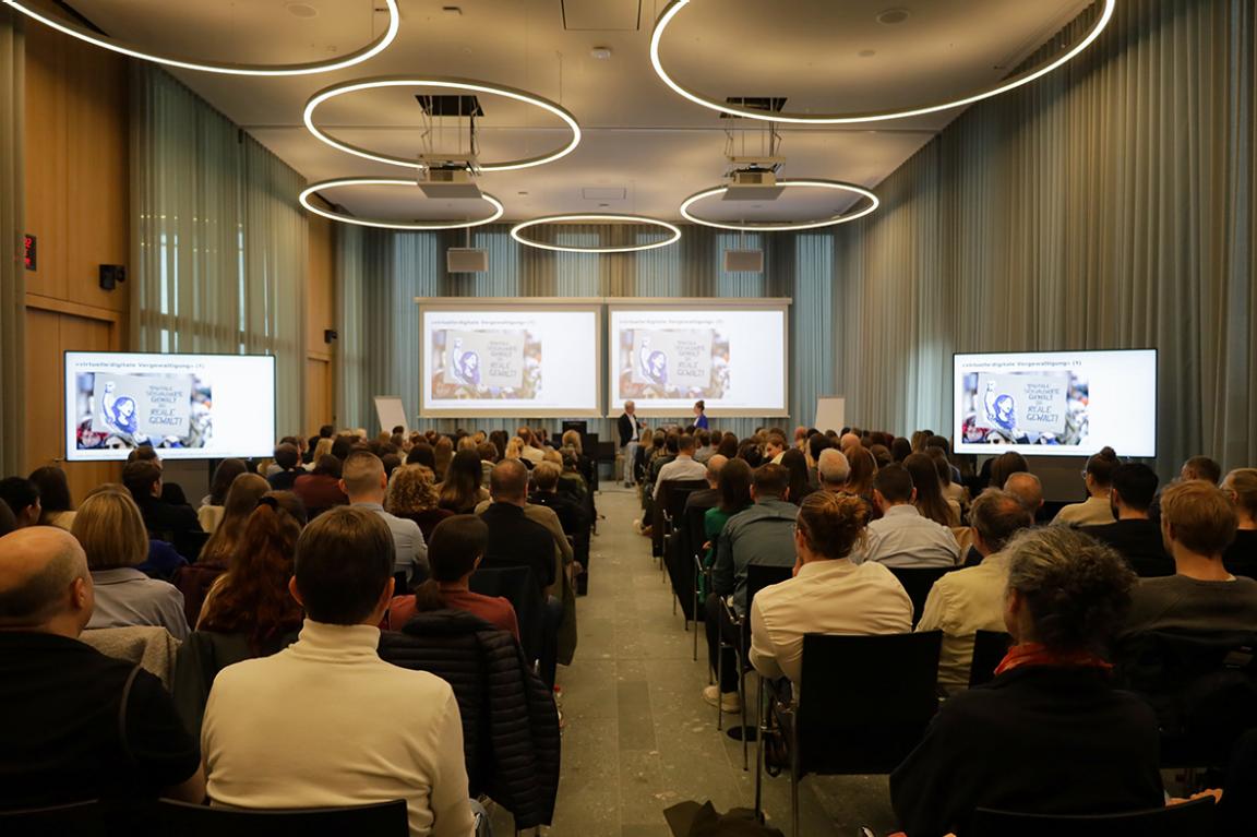 Blick von hinten in einen voll besetzten Veranstaltungssaal mit einer Präsentation auf mehreren Bildschirmen.