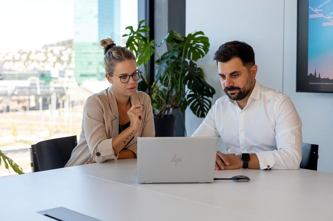 Eine Mitarbeiterin und ein Mitarbeiter sitzen an einem Besprechungstisch und schauen gemeinsam auf einen Laptop.