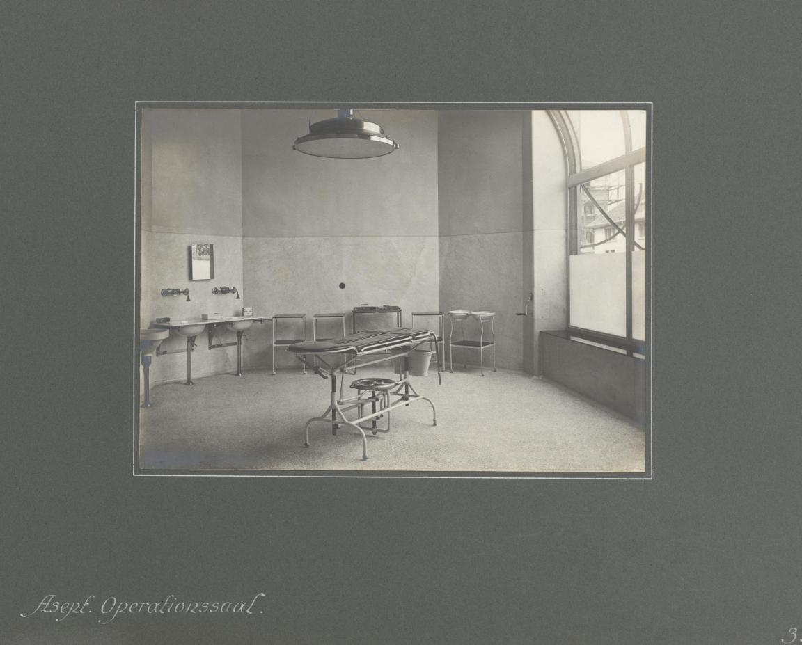 Foto des Operationssaals mit Operationstisch in der Mitte, Lampe darüber, Lavabos und weiteren Utensilien an der Wand