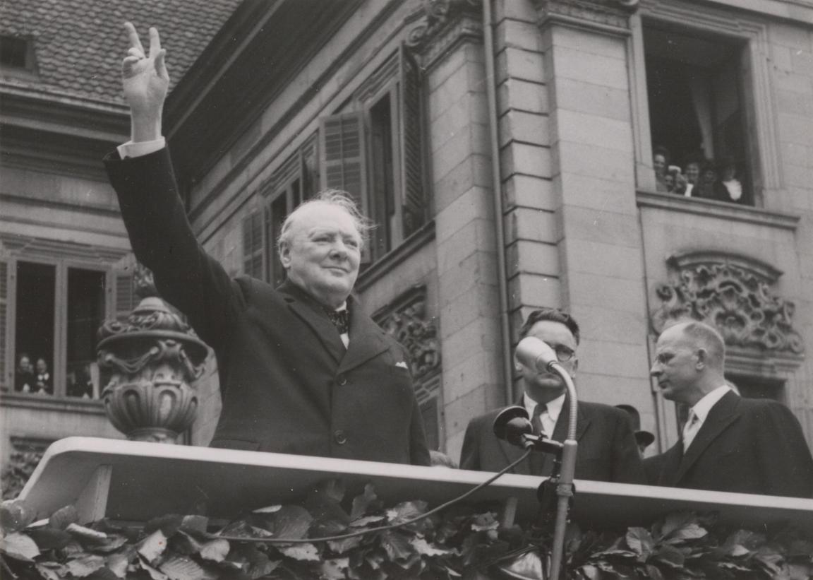 Churchill blickt von der Rednertribüne und macht mit den Fingern der erhobenen rechten Hand das Victory-Zeichen