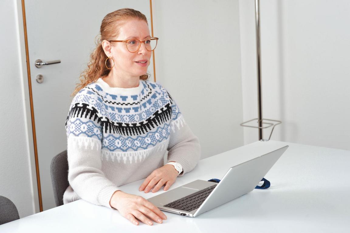 Das Bild zeigt eine sitzende Frau mit Laptop vor sich, die im Gespräch mit einer nicht angezeigten Person ist