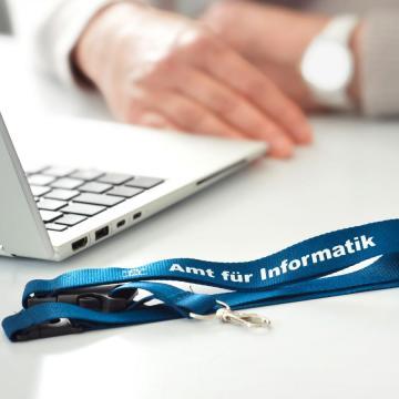 Das Bild zeigt einen Laptop, einen Schlüsselanhänger mit der Beschriftung Amt für Informatik, eine Wasserflasche und die Hände einer Person, die auf der Tischplatte angelehnt sind.