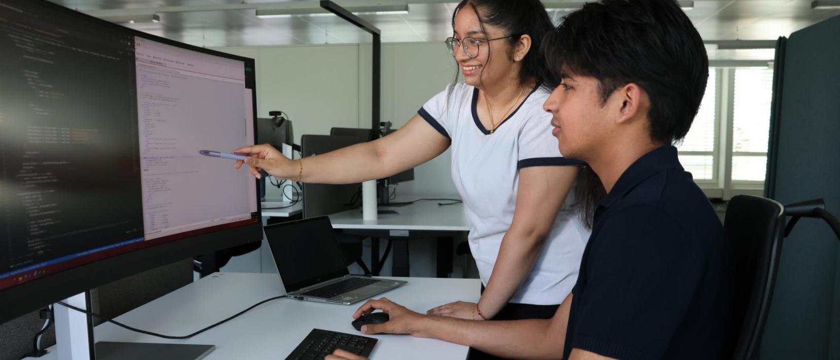 Das Bild zeigt eine stehende, junge Frau, die mit einem Stift auf einen PC-Bildschirm zeigt, neben ihr sitzt ein junger Mann, der seine Hände an der Tastatur und Maus hat und in den PC-Bildschirm schaut.