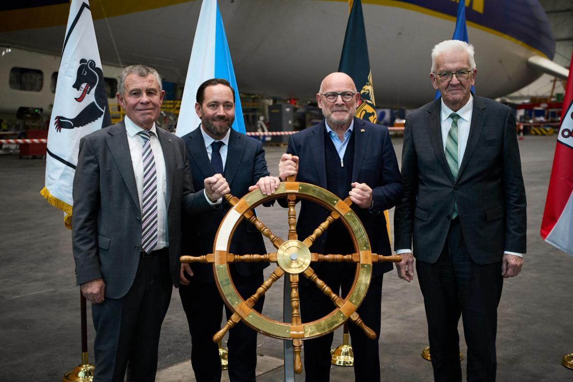 Regierungsrat Ernst Stocker und Ministerpräsident Winfried Kretschmann stehen in einer Zeppelin-Halle um das Steuerrad, das den Vorsitz der Internationalen Bodenseekonferenz symbolisiert 