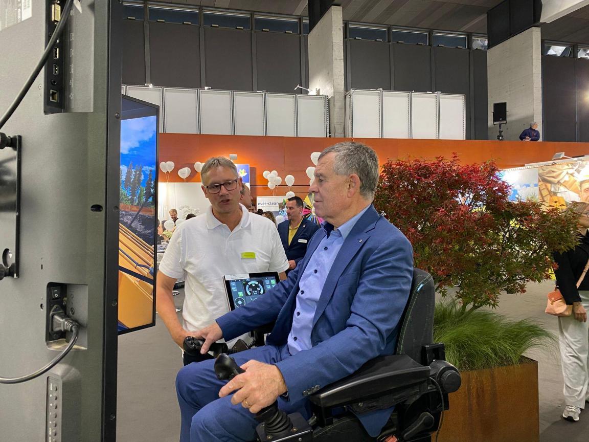 Finanzdirektor Ernst Stocker sitzt in einem Baumaschinensessel vor einem Bildschirm und steuert mit zwei Joysticks einen virtuellen Bagger.